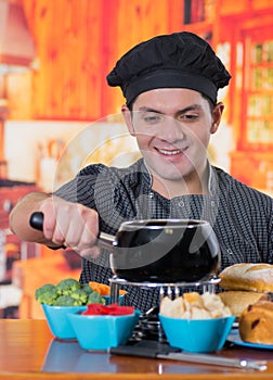 Handsome smiling young cheff preparing a gourmet Swiss fondue dinner with assorted cheeses and a heated pot of cheese