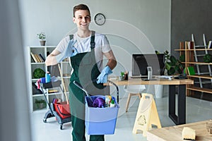 handsome smiling young cleaner holding bucket with cleaning supplies and showing