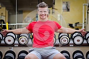 Handsome smiling young man in gym sitting on dumbbells rack