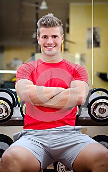Handsome smiling young man in gym sitting on dumbbells rack