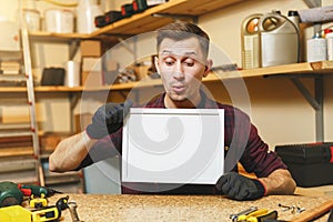 Handsome smiling young man working in carpentry workshop at wooden table place with piece of wood