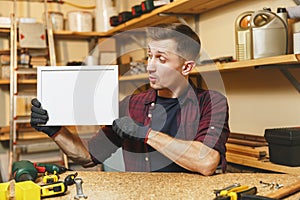 Handsome smiling young man working in carpentry workshop at wooden table place with piece of wood