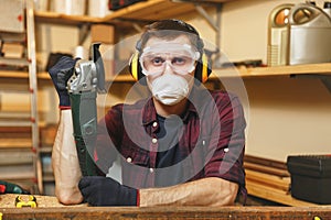 Handsome smiling young man working in carpentry workshop at wooden table place with piece of wood