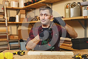 Handsome smiling young man working in carpentry workshop at wooden table place with piece of wood