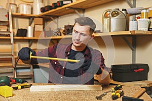 Handsome smiling young man working in carpentry workshop at wooden table place with piece of wood