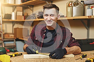 Handsome smiling young man working in carpentry workshop at wooden table place with piece of wood