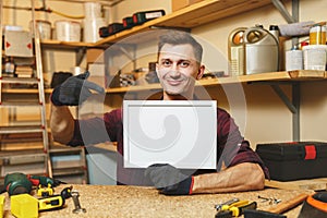 Handsome smiling young man working in carpentry workshop at wooden table place with piece of wood