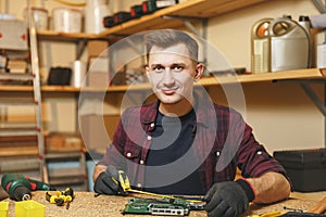 Handsome smiling young man working in carpentry workshop at wooden table place with piece of wood