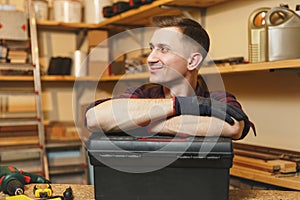 Handsome smiling young man working in carpentry workshop at wooden table place with piece of wood