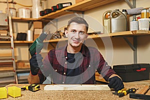 Handsome smiling young man working in carpentry workshop at wooden table place with piece of wood