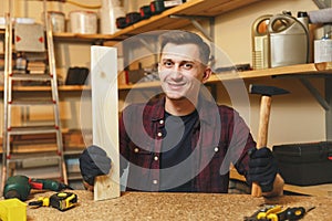 Handsome smiling young man working in carpentry workshop at wooden table place with piece of wood