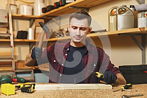 Handsome smiling young man working in carpentry workshop at wooden table place with piece of wood