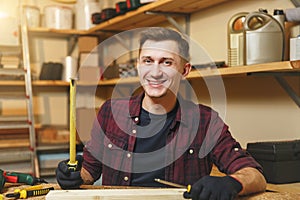 Handsome smiling young man working in carpentry workshop at wooden table place with piece of wood