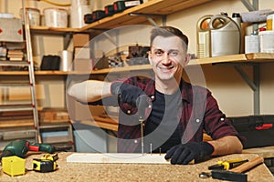 Handsome smiling young man working in carpentry workshop at wooden table place with piece of wood