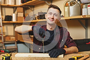 Handsome smiling young man working in carpentry workshop at wooden table place with piece of wood