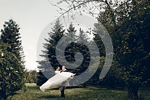 Handsome strong groom holding happy bride in the air, newlywed couple posing in summer park, groom carrying beautiful bride,