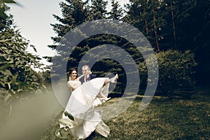 Handsome strong groom holding happy bride in the air, newlywed couple posing in summer park, groom carrying beautiful bride,