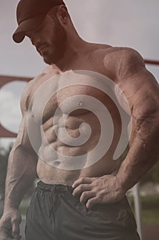 Handsome strong young man with beard wearing black cap with perfect abdominal muscles standing outdoor gym training workout