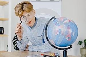 Handsome student is sitting at a desk with magnifying glass in hand. Male is watching to the globe in class