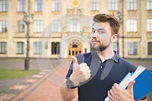 Handsome student standing with books in the hands of the university building background and showing a thumbs up photo