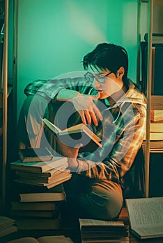 A handsome student wearing glasses, sitting amidst stacks of books within the shelves of a library late in the evening, engrossed