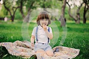 handsome stylish 3 years old toddler child boy with funny face in suspenders enjoying sweets on picnic in spring or summer garden