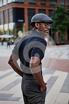 Handsome stylish African American man stands on the street in fashionable attire, wearing a cap, sunglasses, and a