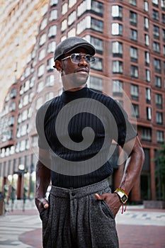 Handsome stylish African American man stands on the street in fashionable attire, wearing a cap, sunglasses, and a