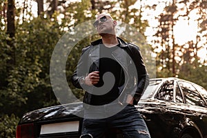 Handsome stylish bald young man in fashion leather jacket in sunglasses stands near modern black automobile outdoors at sunset.