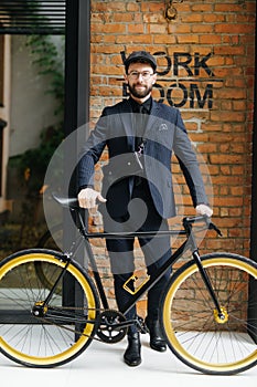 Handsome stylish bearded male wearing suit and hat standing with his bicycle by a brick wall