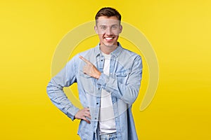 Handsome stylish blond adult man with white smile, pointing finger upper left corner over yellow background, introduce