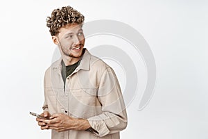 Handsome stylish blond man holding mobile phone, looking behind himself and smiling, reading promo text on copy space