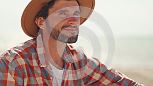 handsome stylish casual man traveler in checkered shirt and hat sitting at sea coastline and enjoying peaceful nature