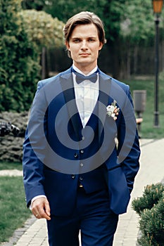 Handsome stylish groom in blue suit walking on a road in the park, newlywed man in elegant clothing posing outdoors, confident