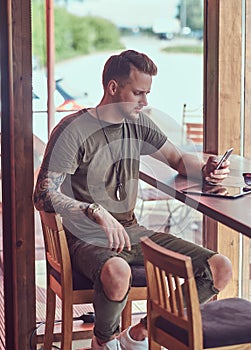 Handsome stylish hipster sits at a table in a roadside cafe, reading the message on the smartphone.