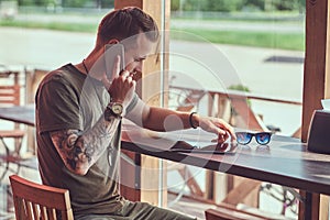 Handsome stylish hipster sits at a table in a roadside cafe, talking on a smartphone.