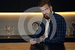 Handsome stylish man in blue suit in cage at home. Sitting in modern black kitchen with smart phone in hand