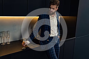 Handsome stylish man in blue suit in cage at home. Standing in modern black kitchen with glass of champagne