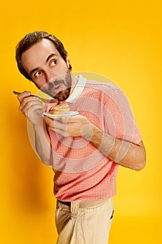 Handsome stylish man with moustache in polo-shirt tasting eclair isolated over yellow background. Facial expression
