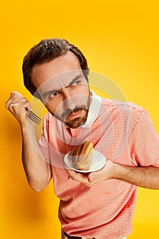 Handsome stylish man with moustache in polo-shirt tasting eclair isolated over yellow background. Facial expression