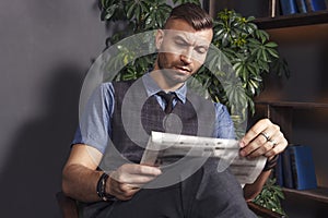 Handsome stylish man sits in chair and reads latest news in newspaper. confident brutal businessman in luxury apartments.