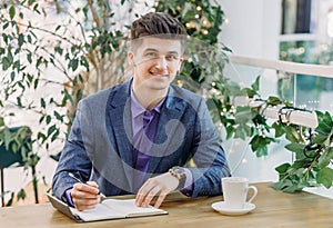 A handsome successful young businessman in a stylish blue suit is sitting in a summer cafe with a pen in his hand and smiling at