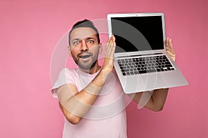 Handsome surprised and amazed man holding laptop computer looking at camera in t-shirt on isolated pink background