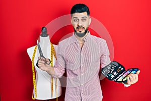 Handsome tailor man with beard holding sew kit and scissors clueless and confused expression