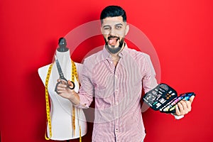 Handsome tailor man with beard holding sew kit and scissors sticking tongue out happy with funny expression