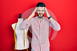 Handsome tailor man with beard standing by manikin with hand on head, headache because stress