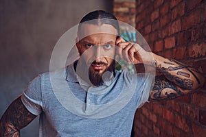 A handsome tattoed male with a stylish haircut and beard, in a gray t-shirt, standing leaning against a brick wall in a