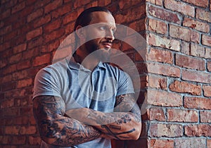 A handsome tattoed male with a stylish haircut and beard, in a gray t-shirt, standing leaning against a brick wall in a