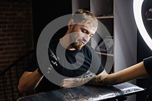 Handsome tattoo master making black tattoo with paint. Master works in black sterile gloves. Young tattoo artist in work