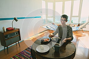 Handsome teen boy eating in vintage room with old fashioned armchair, retro radio turntable, table with food, telephone, standart